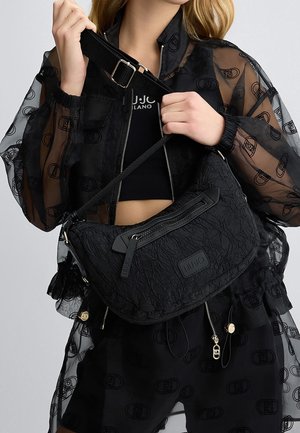 Femme portant une veste noire transparente à motifs et un top court, tenant un sac à bandoulière noir texturé avec fermeture éclair et patch de marque.