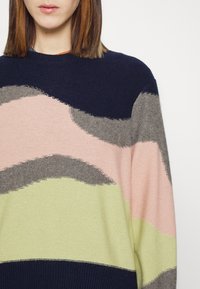 Femme portant un pull à blocs de couleur avec des bandes ondulées bleu marine, rose, grises et vert clair, montrée des épaules jusqu'au milieu du torse.