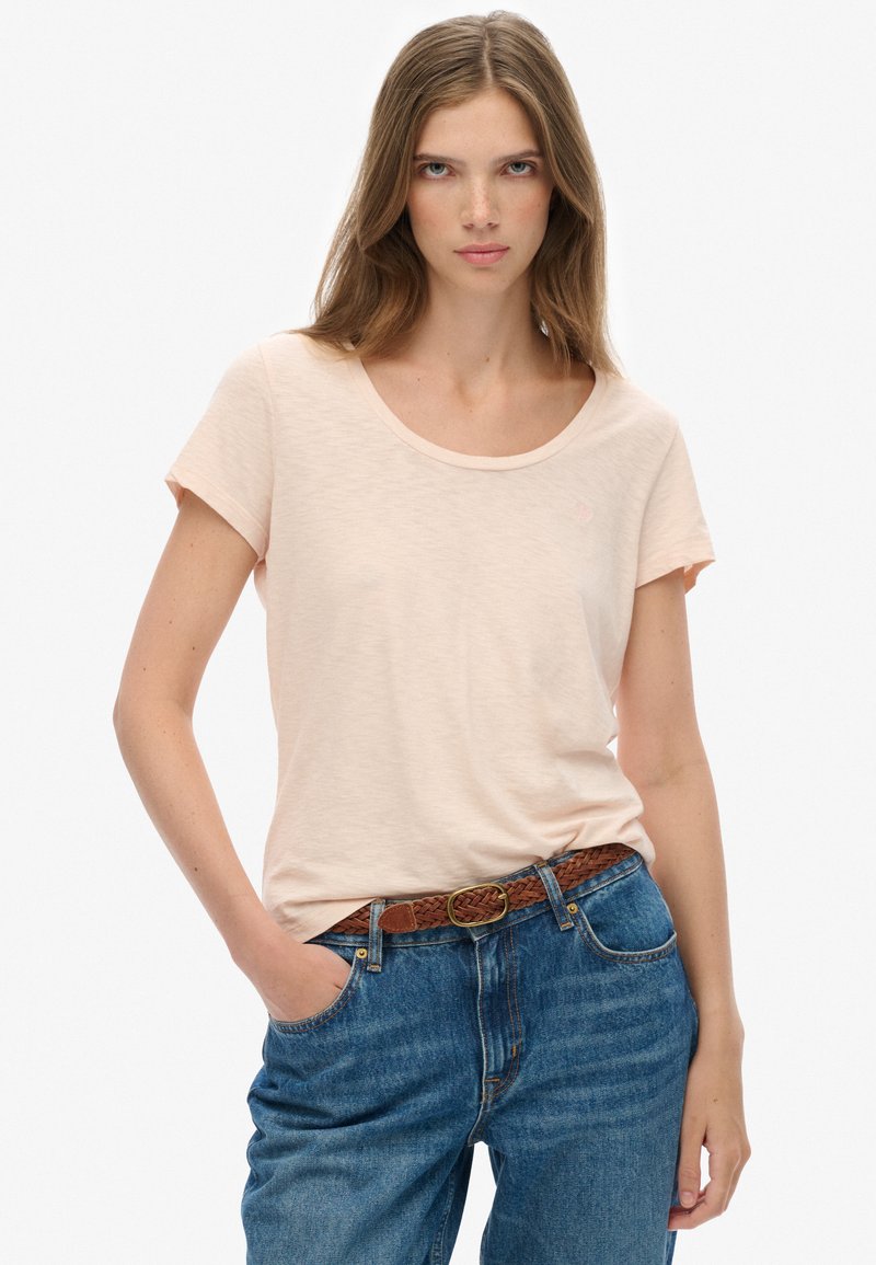 Camiseta de manga corta en color durazno claro con cuello redondo, hecha de un tejido suave, combinada con jeans de denim azul de cintura alta y un cinturón trenzado marrón.