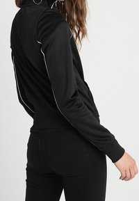 Chaqueta atlética negra con un cuello alto, con ribetes blancos a lo largo de las mangas, hecha de una tela suave y elástica.