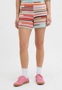 Persona vistiendo pantalones cortos de punto a rayas multicolores, calcetines blancos hasta la altura del tobillo y zapatillas rosas con cordones, de pie sobre un fondo blanco liso.