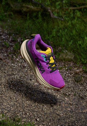 Scarpa da corsa viola con una tomaia in rete traspirante, dettagli gialli, lacci neri e una suola spessa e testurizzata con dettagli rossi, sospesa sopra ghiaia.