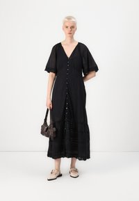Robe maxi noire avec un décolleté en V, des manches courtes en dentelle, une fermeture à boutons à l'avant et un ourlet superposé. Assortie à un petit sac marron et des chaussures beiges.