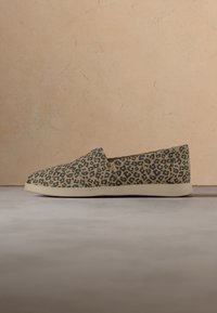 Chaussure slip-on en tissu beige à imprimé léopard noir avec une semelle plate blanc cassé sur un fond neutre.