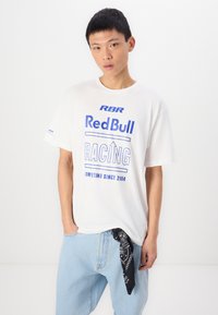 T-shirt en coton blanc avec impression graphique bleue avec le texte "RBR Red Bull RACING Competing Since 2004." Associé à un jean bleu clair et un bandana noir.