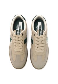 Zapatillas beige con acentos verdes y ante texturizado. Presentan cordones gruesos y suelas de goma. El interior tiene una plantilla con marca. Diseño atlético clásico.