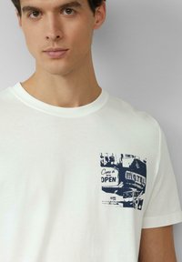 Witte katoenen t-shirt met een marineblauw grafisch print van een vintage motelbord met de tekst "Kom binnen, we zijn OPEN" op de linkerborst.