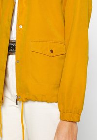 Żółta kurtka typu bomber wykonana z lekkiego materiału, wyposażona w zamek błyskawiczny, elastyczne mankiety oraz kieszeń na piersi zapinaną na napy.