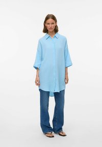 Light blue button-up shirt in textured fabric with a collar and loose fit. Worn with blue jeans and flip-flops.
