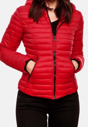 Donna che indossa una giacca trapuntata rossa con tasche con cerniera, pantaloni neri e mani nelle tasche, mostrata dalle spalle fino ai fianchi.