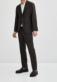 Un uomo indossa un completo marrone su misura con una camicia bianca a colletto, abbinato a mocassini neri; lo sfondo neutro mette in risalto il suo abbigliamento.