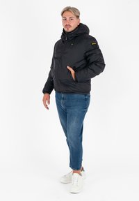 Zwarte gewatteerde jas met een capuchon, voorzien van een ritssluiting en twee zijzakken. Gekocht met blauwe jeans en witte sneakers. Glad materiaal.