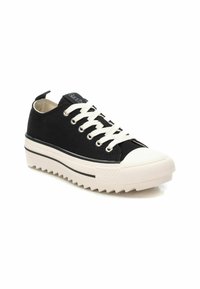 Zapatilla de lona negra con cordones blancos y puntera de goma. Presenta una suela de plataforma blanca gruesa con un patrón en zigzag y una sutil raya negra.