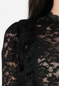 Musta pitsipaita, jossa on monimutkaisia kukkakuvioita, röyhelöhihoja ja läpikuultava rakenne, luoden elegantin kerrosmaisen ilmeen.