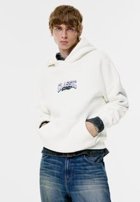 Witte capuchontrui met "CLASSIC" in kleurrijke tekst geborduurd, gedragen over een donker denim shirt, gecombineerd met een blauwe spijkerbroek.