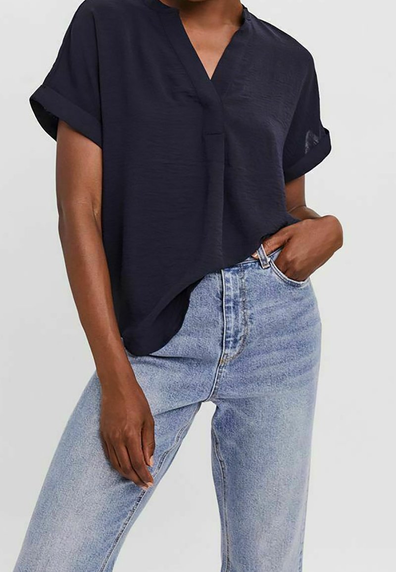 Femme portant un chemisier ample à manches courtes bleu marine foncé avec des poignets roulés, rentré dans un jean taille haute bleu clair, une main dans la poche.