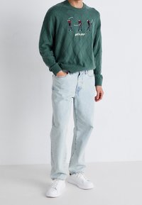 Pull vert tricoté avec des motifs en losange, orné de broderies sur le thème du golf, associé à un jean bleu clair et des baskets blanches.