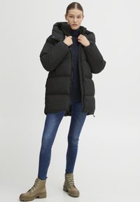 Zwarte gewatteerde jas met een afneembare capuchon, ritssluiting en geribde manchetten, gecombineerd met blauwe jeans en tan geknoopte laarzen.