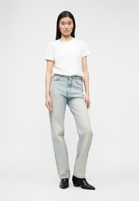 Γυναίκα που στέκεται, φοράει λευκό κοντομάνικο πουκάμισο, ανοιχτό μπλε ψηλόμεσα τζιν και μαύρα μποτάκια με τακούνι.