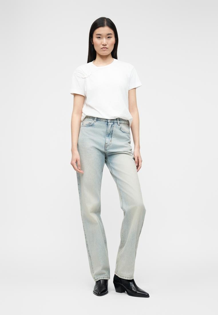 Femme debout portant une chemise blanche à manches courtes, un jean taille haute bleu clair, et des bottines à talons noires.