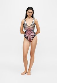 Femme debout pieds nus, portant un maillot de bain une pièce à rayures géométriques noires, blanches et roses, sur fond blanc uni.