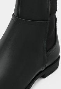 Čierna členková členková topánka z hladkého syntetického materiálu, s elastickými bočnými panelmi a plochou podrážkou. Má zaoblenú špičku a minimálne švy.