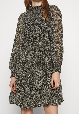 Femme portant une robe noire à manches longues avec motif floral, un col haut froncé et des poignets, ainsi qu'une jupe fluide mi-longue au genou.