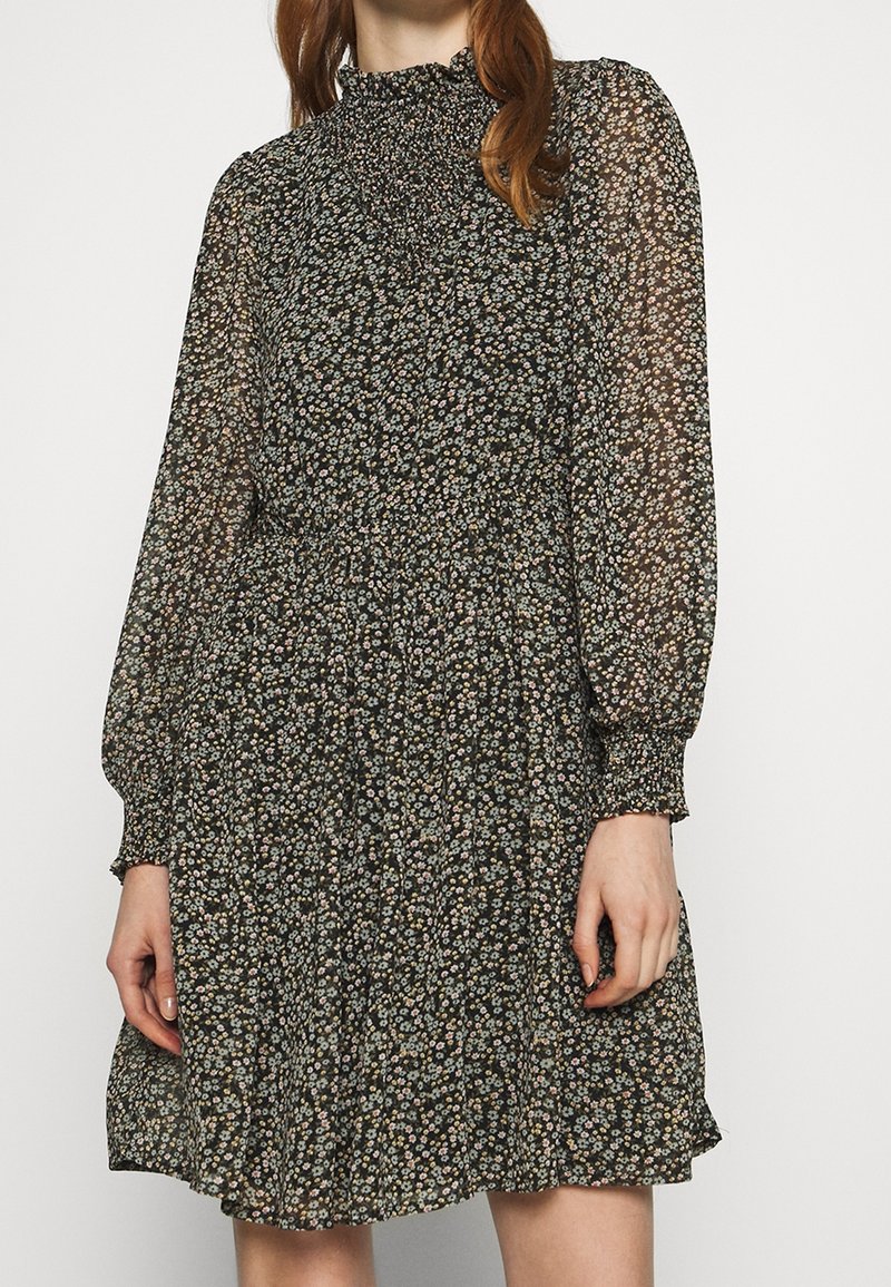 Femme portant une robe noire à manches longues avec motif floral, un col haut froncé et des poignets, ainsi qu'une jupe fluide mi-longue au genou.