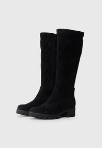 Paire de bottes hautes jusqu'au genou en daim noir avec semelles crantées épaisses et fermetures éclair latérales, debout sur un fond gris clair.