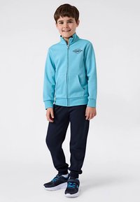 Jasnoniebieska bluza z kapturem na zamek z kieszeniami, czarnym logo, zestawiona z granatowymi dresami i czarnymi tenisówkami z niebieskimi akcentami.