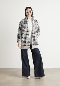Graham- und schwarze Hahnentritt-Musterjacke mit Reißverschluss, hochgeschlossenem Kragen und beigen Akzenten, kombiniert mit dunklen weit geschnittenen Jeans und weißen Absätzen.