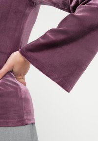 Fløyelstopp i malva med vide ermer, som har en jevn tekstur og enkel kant, fremhevet av en innsvingt silhuett ved midjen.