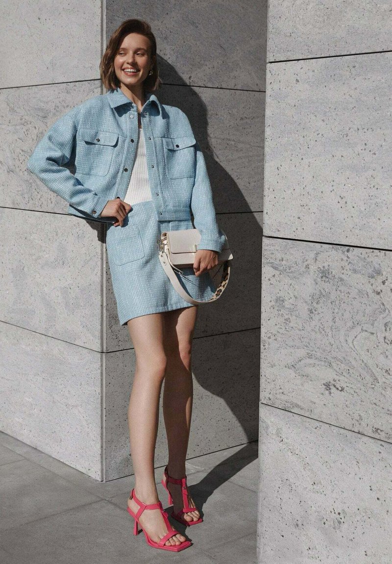 Lichtblauwe, gestructureerde jas en bijpassende rok met twee zakken. Gecombineerd met een witte top en roze hoge sandalen. Vierkante, neutrale handtas.