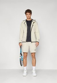 Heledat beeži lukuga jope mustade aktsentidega, must t-särk, beežid lühikesed püksid, valged tossud, tennisereket käes.