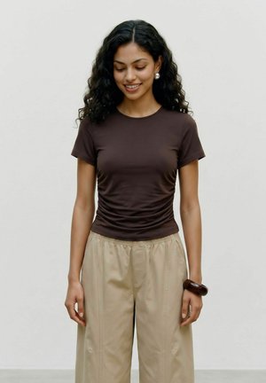 Jeune femme aux cheveux noirs bouclés porte un t-shirt marron ajusté, un pantalon beige à jambes larges, des boucles d'oreilles en perles et un bracelet foncé, souriante et regardant vers le bas.