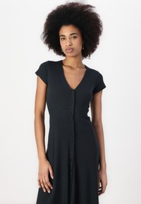 Schwarzes, tailliertes Kleid mit kurzen Ärmeln, tiefem V-Ausschnitt und einer sechs Knopf-Leiste vorne; aus geripptem Material mit glatter Textur gefertigt.
