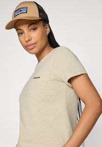 Mulher com cabelo trançado, usando um boné beige da Patagonia e uma t-shirt beige da Patagonia combinando, olhando ligeiramente para o lado contra um fundo branco.