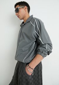 Veste en tissu imperméable grise avec des bandes blanches, des poignets élastiques et une fermeture éclair à l'avant ; associée à un pantalon de survêtement à carreaux noirs et des accessoires argentés.