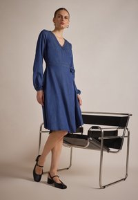 Denim omslagskjole i dyp blå, med lange puff ermer og V-hals. Kombinert med svarte, chunky hæler og en moderne stol.