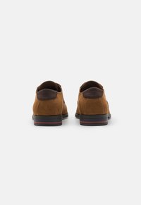Pantofi din suede maro cu o secțiune din spate netedă și un profil jos. Prezintă o talpă neagră cu o bandă accent roșie. Design clasic, fără șireturi.