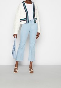 Valkoinen palmikkoneulottu lyhyt neuletakki monivärisillä raidallisilla reunoilla, yhdistetty vaaleansinisiin liehuviin farkkuihin ja valkoisiin remmikenkiin. Sininen käsilaukku.