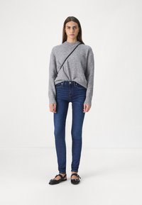 Grå strikket genser med lange ermer, kombinert med mørkeblå tettsittende jeans og svarte flate sko, med en tynn crossover-stropp.