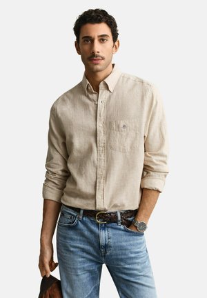 Uomo con baffi che indossa una camicia beige con bottoni, jeans blu, cintura intrecciata marrone, orologio da polso argentato, tiene un cappello marrone nella mano sinistra.