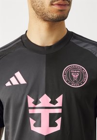 Crna sportska majica s kratkim rukavima, s ružičastim detaljima uključujući Adidasov logo i okrugli amblem za Klub Internacional de Fútbol Miami.