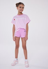 Camiseta rosa con el logo de Champion y mangas cortas, combinada con pantalones cortos rosa claro. Zapatillas blancas y rosas completan el look deportivo.