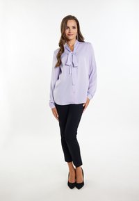 Levendula színű blúz, nyakán masnival, hosszú ujjú, fekete, slim-fit nadrággal és fekete magassarkú cipővel párosítva. Egyszerű háttér.