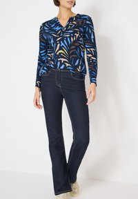 Bluzka z długim rękawem i dekoltem w serek z abstrakcyjnym wzorem w kolorach niebieskim, beżowym i zielonym; zestawiona z ciemnymi, rozkloszowanymi dżinsami i jasnymi trampkami.