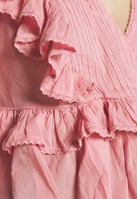Růžové bavlněné šaty s volánovými rukávy, skladovanou texturou a jemným krajkovým lemem podél okrajů, zdůrazňující vrstvený design.