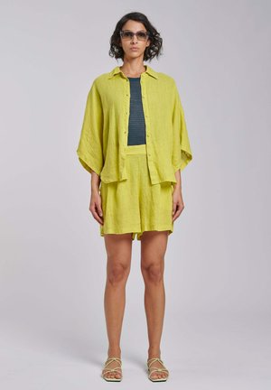 Kobieta z krótkimi, kręconymi włosami, nosząca duże kwadratowe okulary przeciwsłoneczne, żółtą koszulę na guziki i zestaw szortów, ciemnoniebieski top oraz żółte sandały, stojąca.