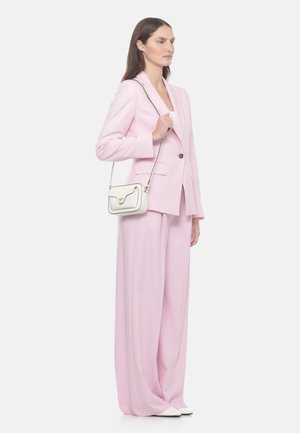 Femme portant un tailleur-pantalon rose clair et des chaussures blanches, tenant un petit sac à bandoulière blanc avec une fermeture dorée, debout devant un fond uni.